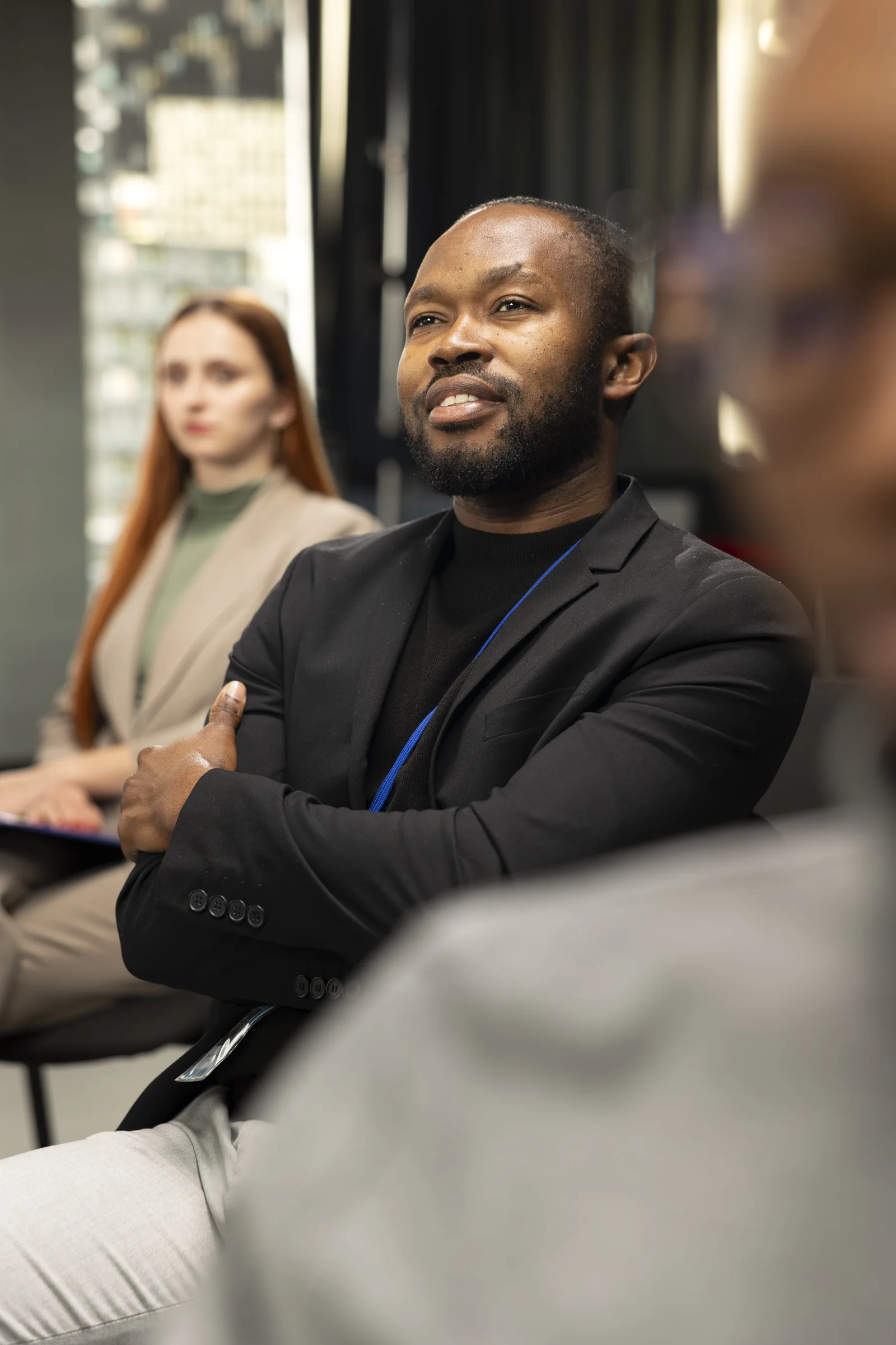 Man in black jacket sitting with arms crossed listening attentively in a meeting.