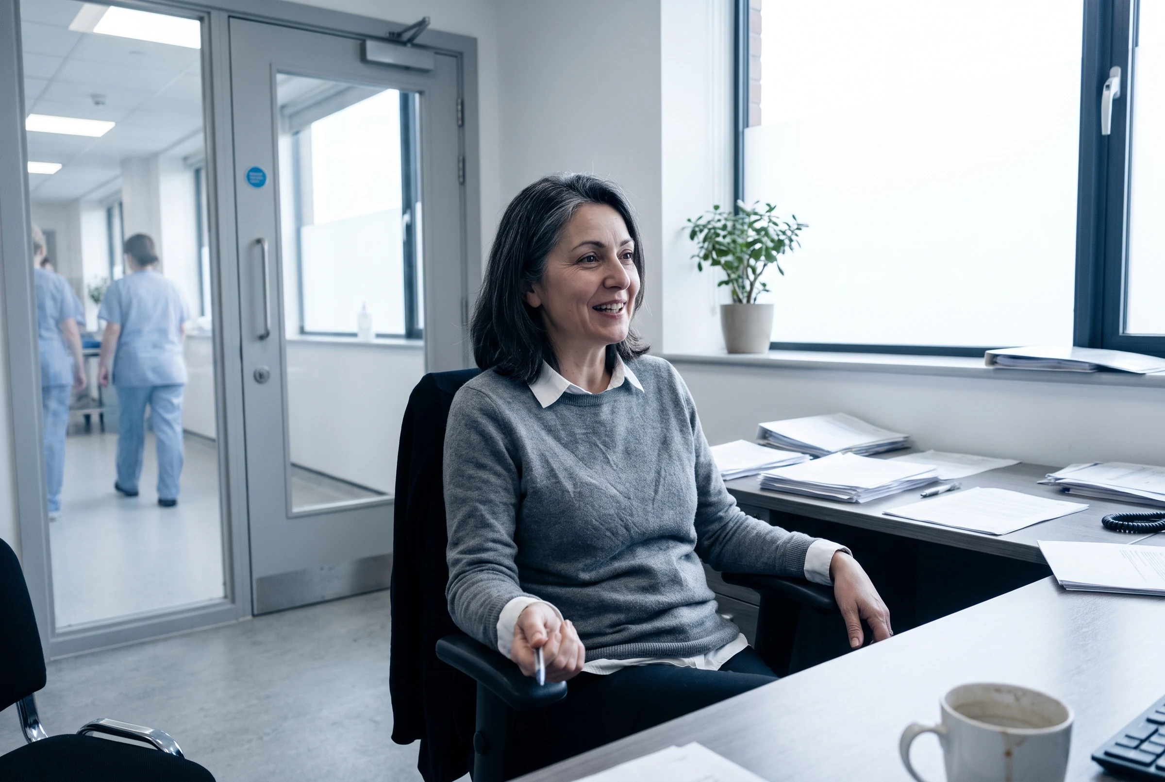 A clinician seated at a desk in a bright hospital office, smiling and looking off camera, with a corridor and colleagues visible through a glass door behind.
