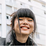 Smiling young woman with straight black hair and bangs, wearing a black jacket outdoors near a building.