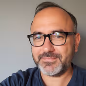Middle-aged man with glasses, short beard, and mustache smiling against a plain gray background.
