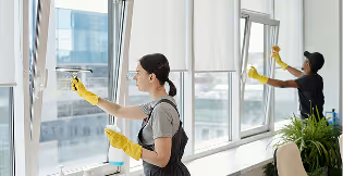 Two people wearing yellow gloves cleaning large office windows with squeegees.