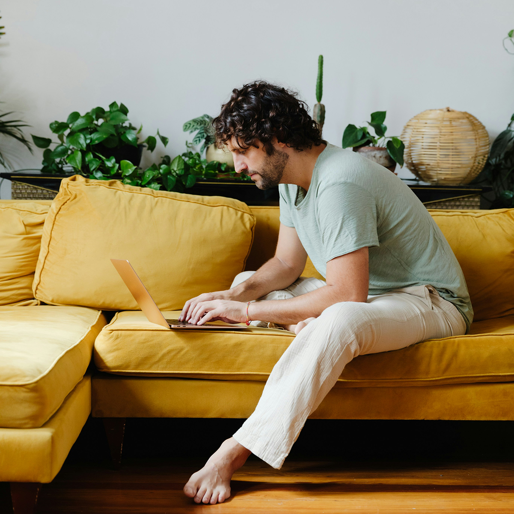 Man with curly hair sitting on a yellow couch, typing on a laptop with plants in the background.