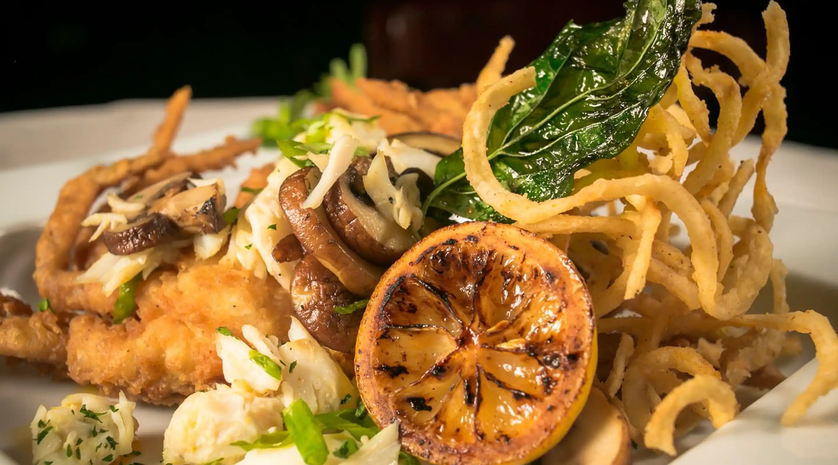 Plate with fried fish topped with sautéed mushrooms, shredded crab, a grilled lemon slice, fried onion strings, and a basil leaf garnish.