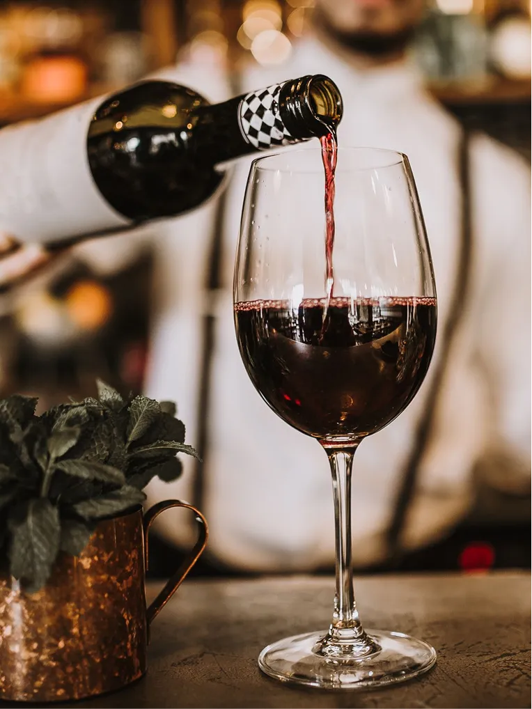 Red wine being poured from a bottle into a wine glass on a table with a copper mug holding green leaves.