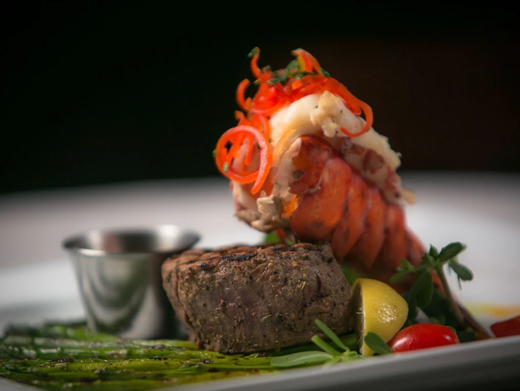 Grilled steak and lobster tail served with asparagus, lemon wedge, cherry tomato, and a small metal sauce cup on a white plate.
