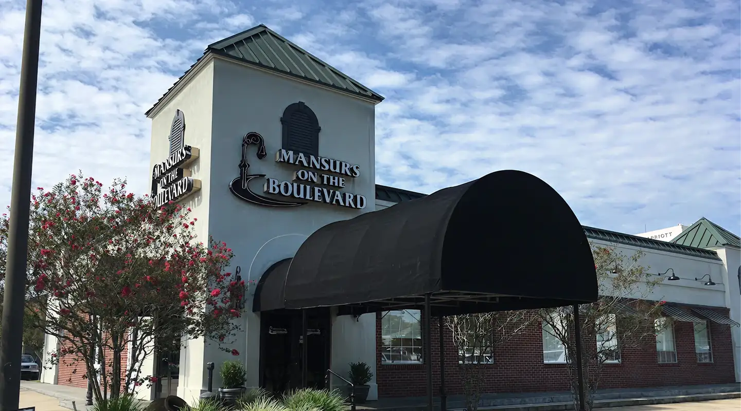 Exterior of Mansurs on the Boulevard restaurant with a black canopy entrance and flowering trees under a partly cloudy sky.