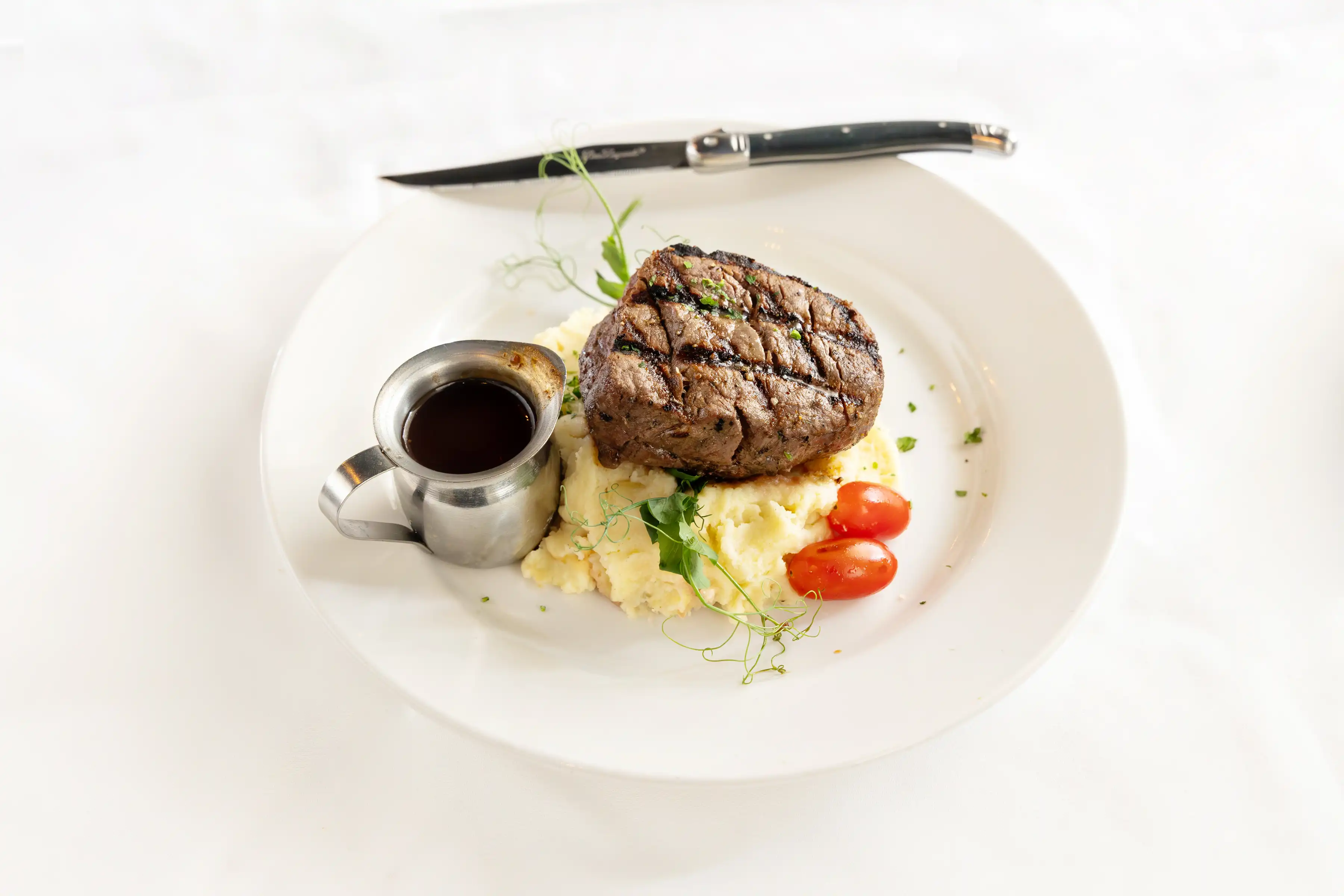 Grilled steak with mashed potatoes, two cherry tomatoes, greens, and a small metal cup of sauce on a white plate with a knife.