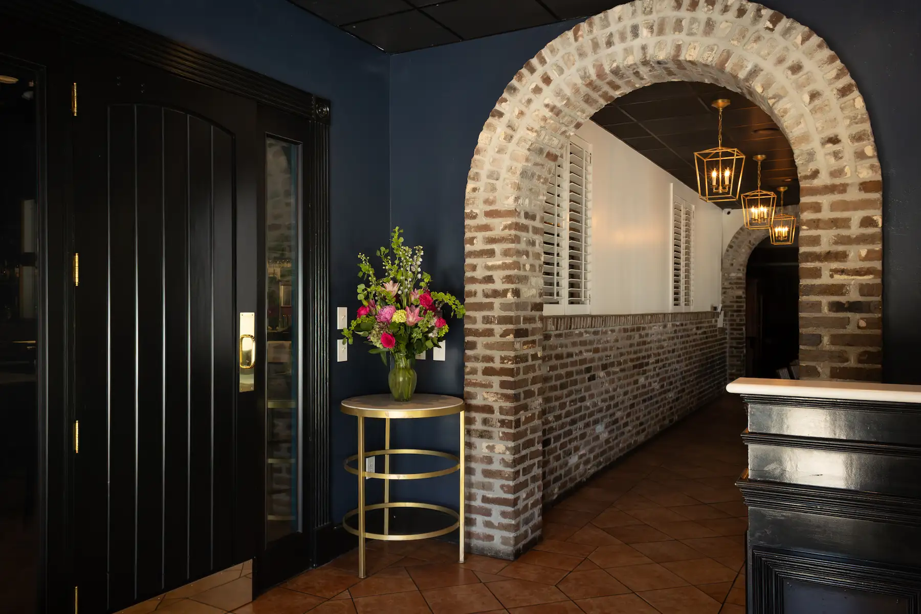 Interior hallway with dark paneled door, brick archways, hanging lanterns, and a round table with a vase of colorful flowers.