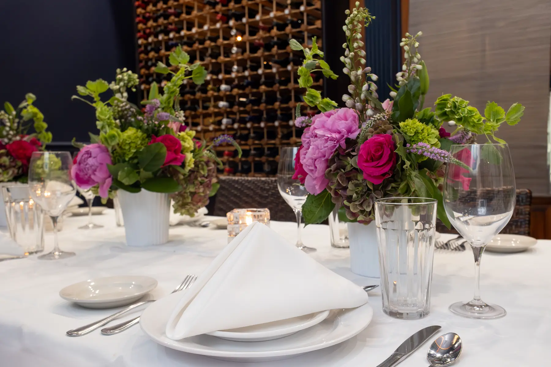 Elegant dining table set with white plates, folded napkins, silver cutlery, glasses, and floral centerpieces featuring pink and red flowers.