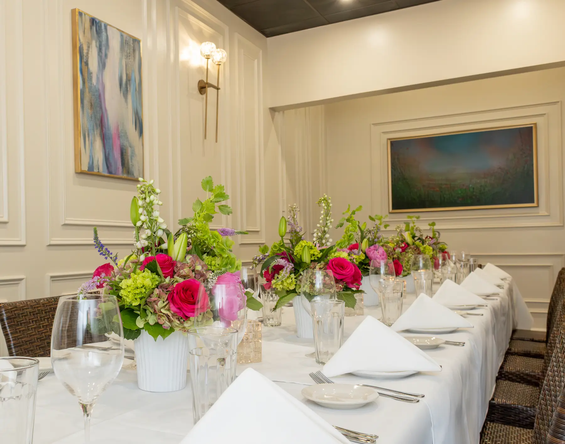 Long dining table with white tablecloth, neatly folded white napkins, glassware, and bouquets of pink and green flowers in white vases, set in an elegant room with framed artwork on the walls.