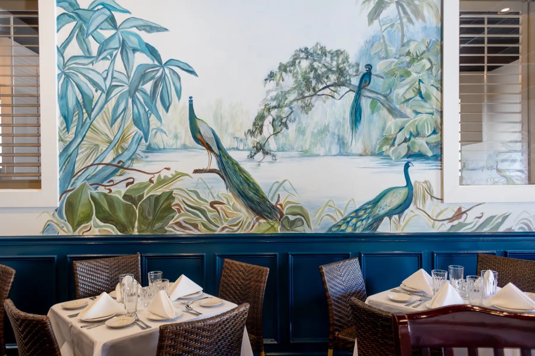 Restaurant dining area with tables set with white napkins and glassware in front of a wall mural featuring peacocks and lush foliage.