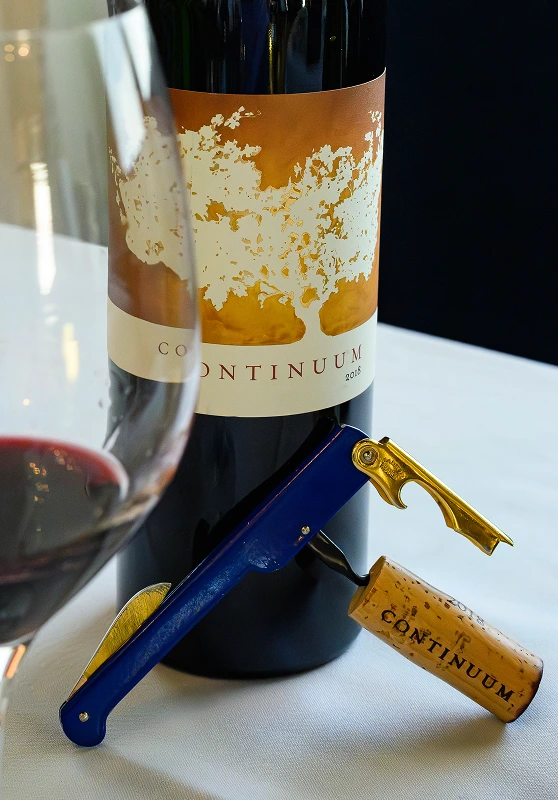 Bottle of Continuum 2018 wine with corkscrew and cork on a white tablecloth, glass of red wine in foreground.