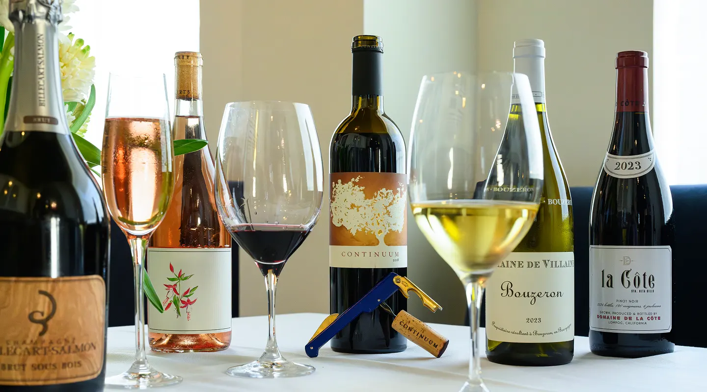 Variety of wine bottles and filled glasses on a white tablecloth, including sparkling, rosé, red, and white wines.