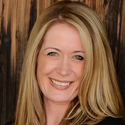 A smiling person with long blonde hair, in front of a dark wood wall.