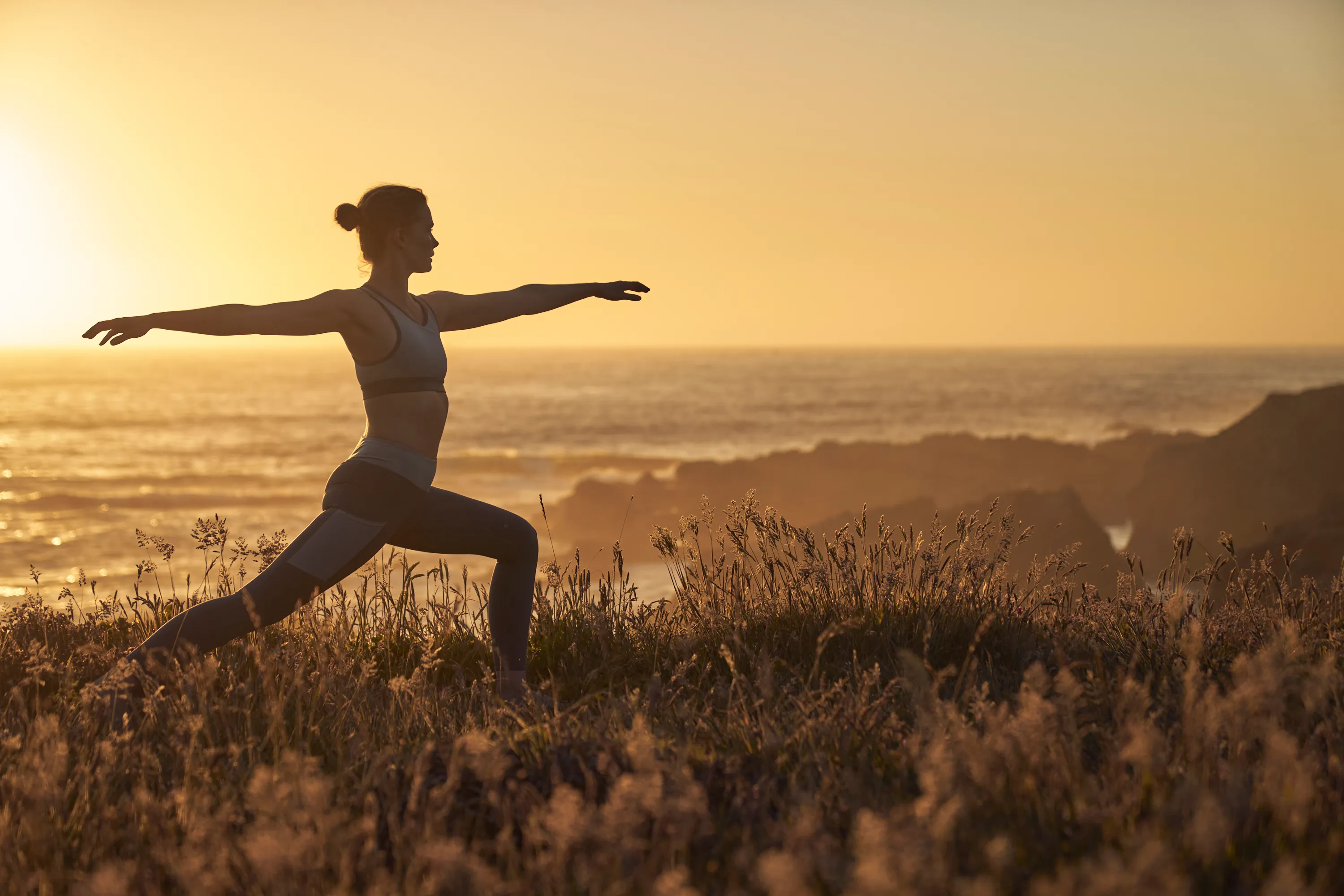 A guest holding a warrior yoga pose on a grassy coastal bluff at sunset.