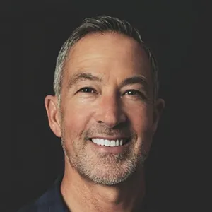 A smiling person with short grey hair and a beard, in a dark shirt against a black background.