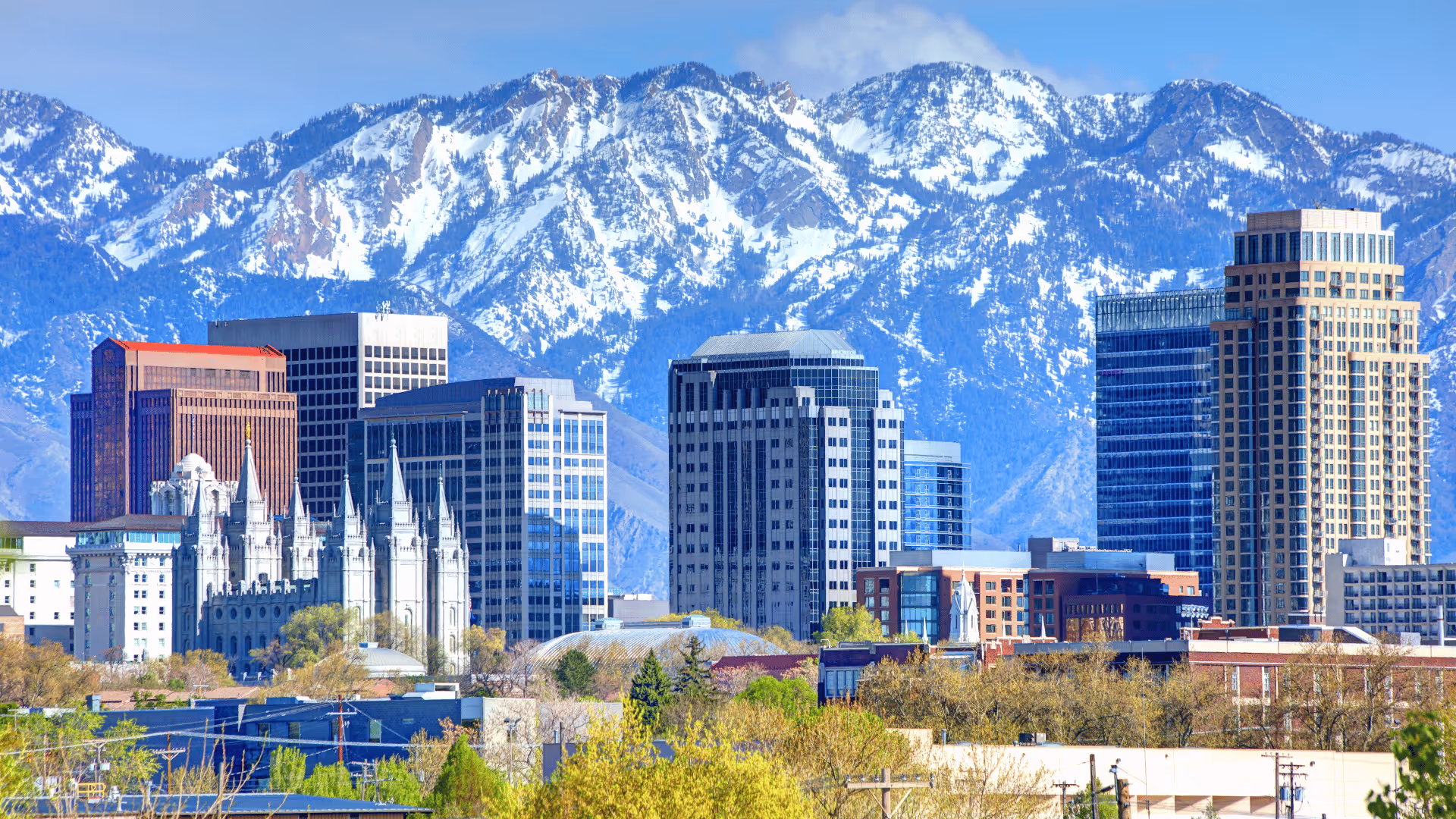 Salt Lake City skyline with modern buildings and historic temple in front of snow-capped mountains.
