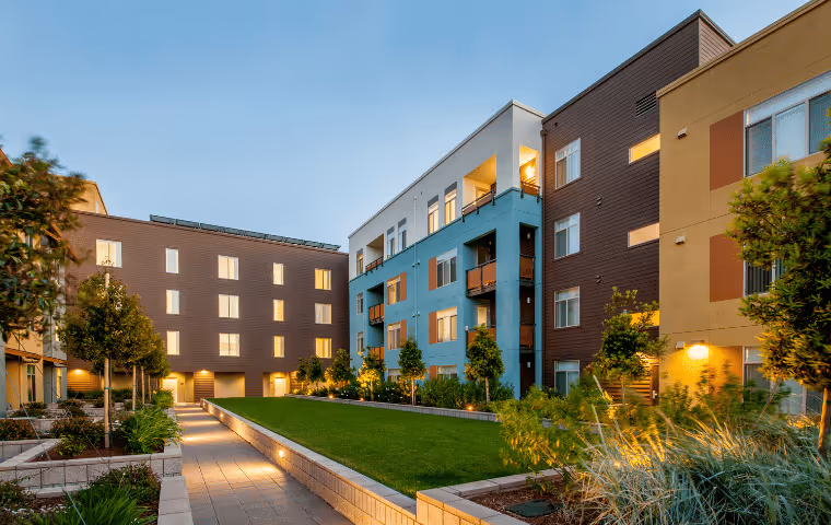 Modern apartment complex at dusk with a well-lit courtyard featuring green lawn, trees, and paved walkways.