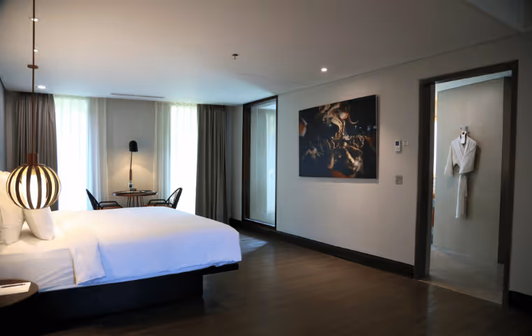 Modern hotel room with a large white bed, round lamp, two chairs and table by window, and a bathrobe hanging in an adjoining bathroom.