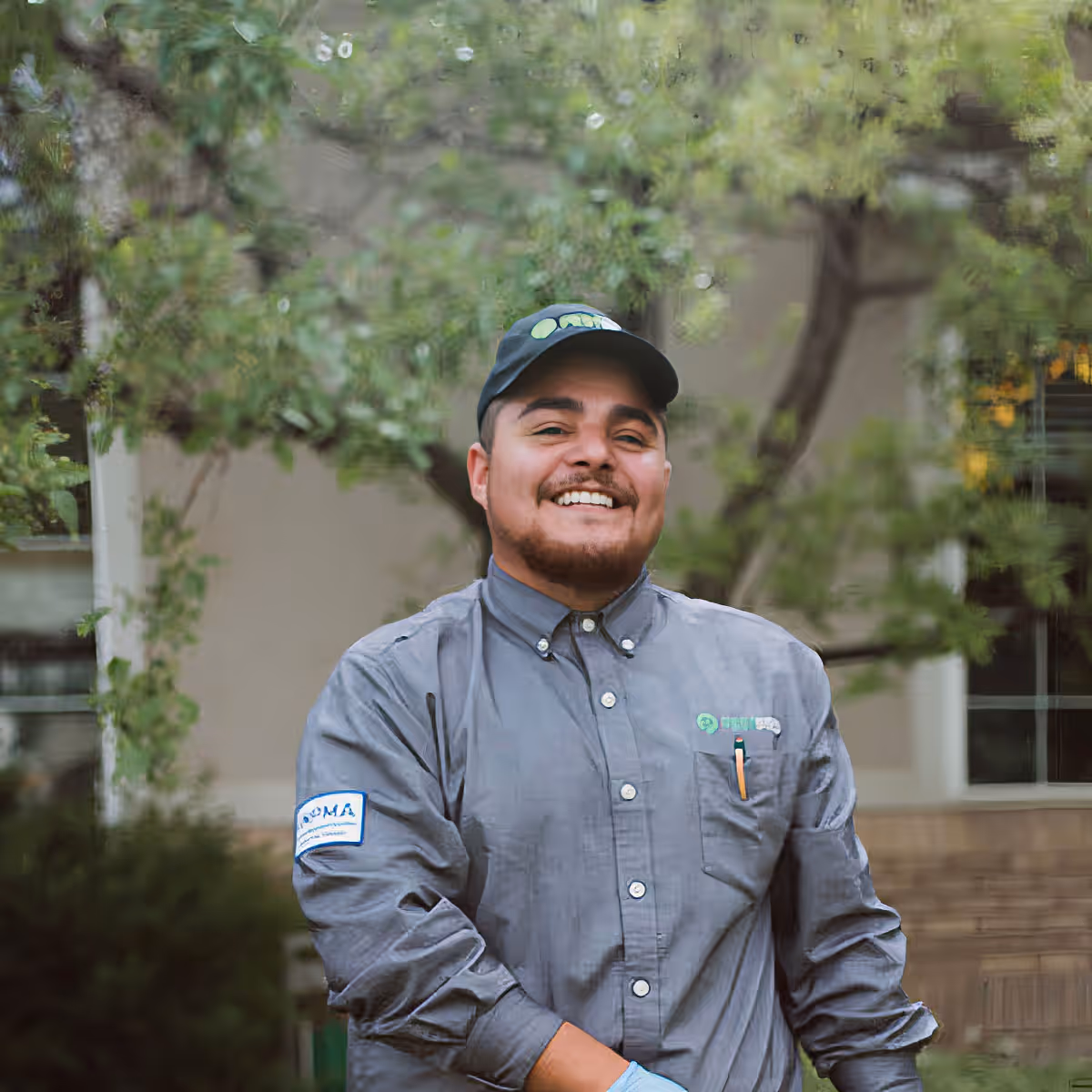 Smiling male pest control technician wearing gray uniform and black cap standing outdoors with trees in the background.