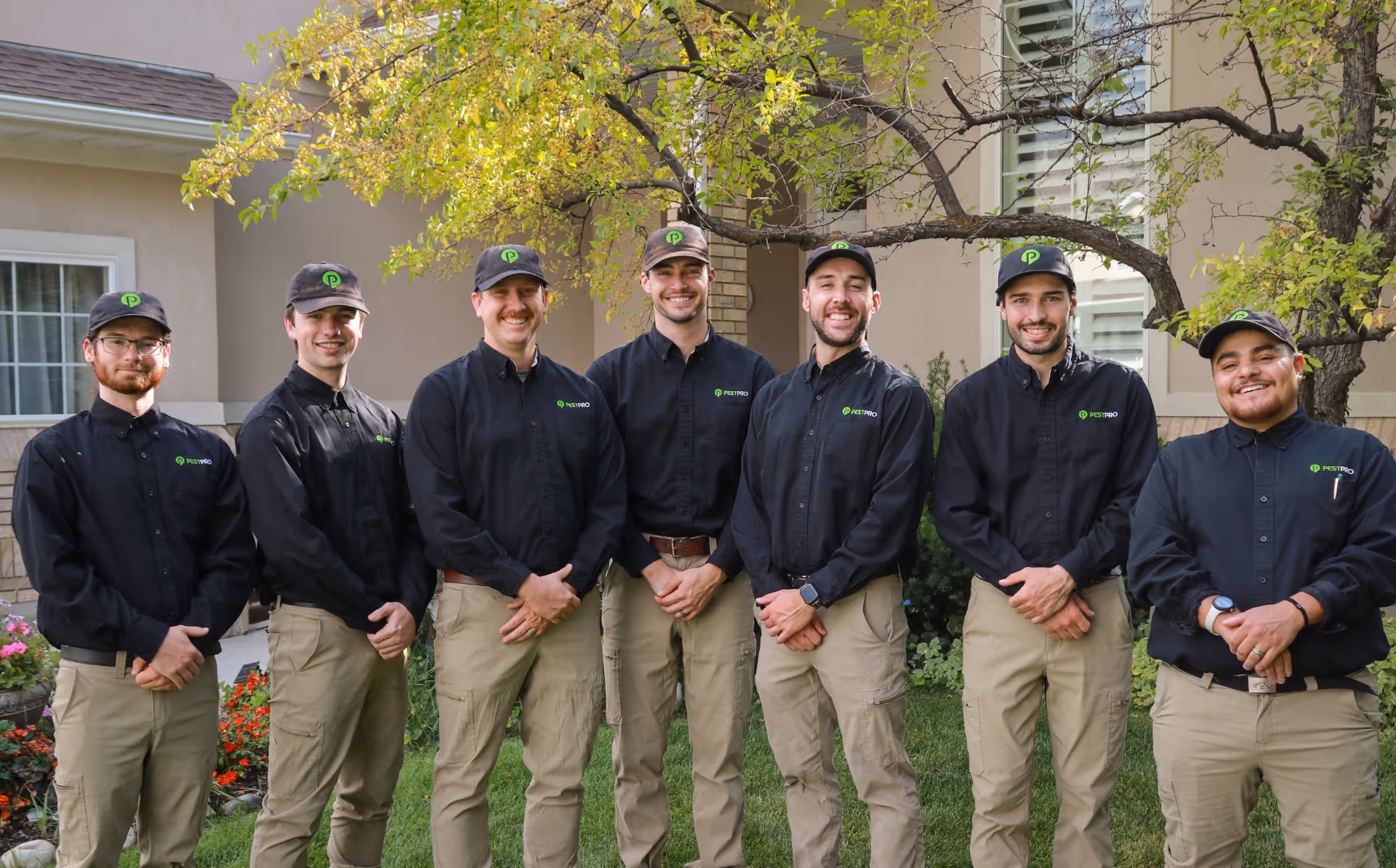 Seven men wearing black Pest Pro shirts, green logo caps, and tan pants standing outdoors in front of a house and garden.