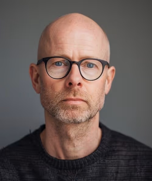 A bald man wearing glasses and a sweater, looking thoughtfully to the side.