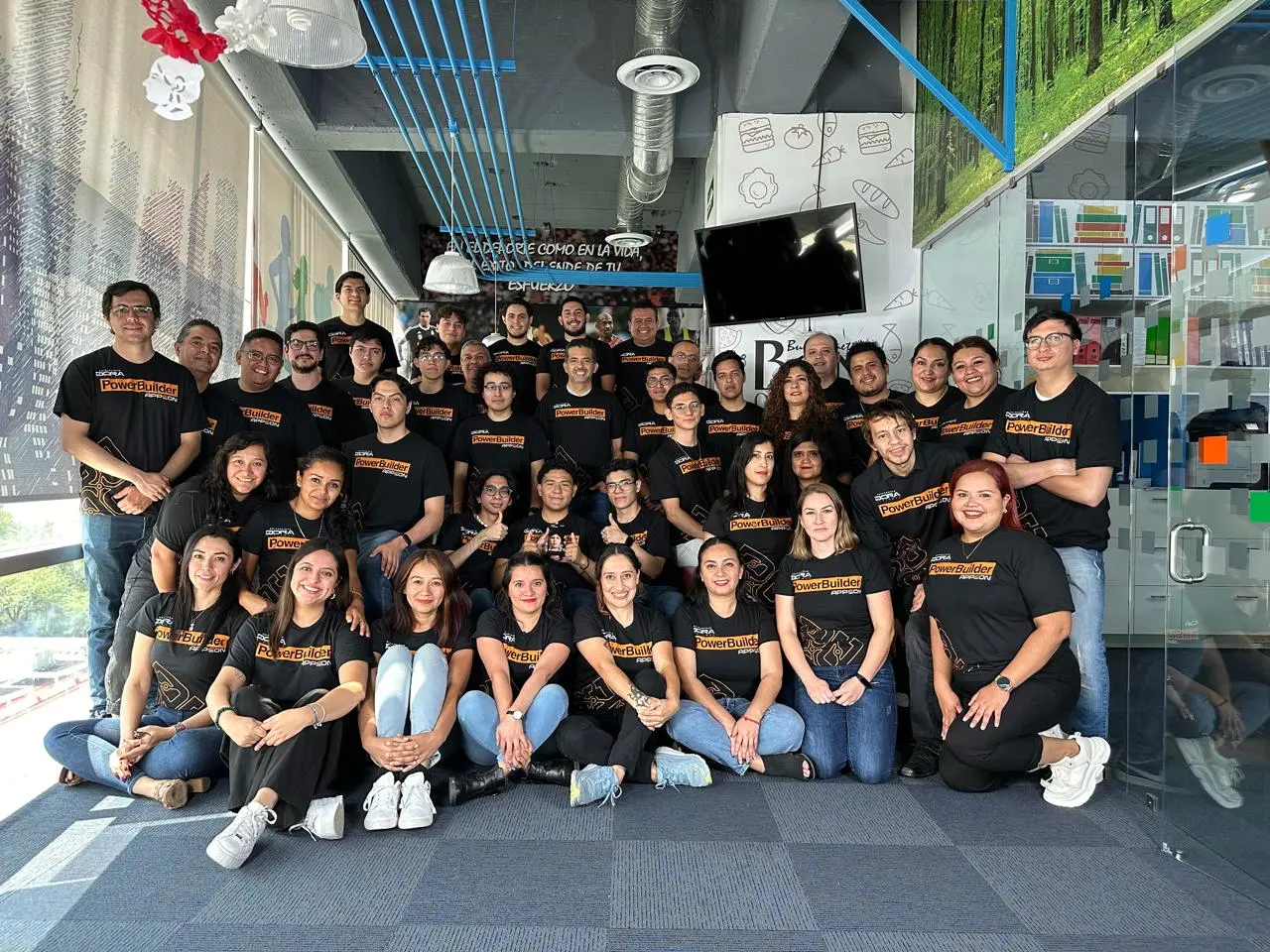 Large group of men and women wearing matching black PowerBuilder t-shirts posing indoors in an office space with glass walls and a TV screen.