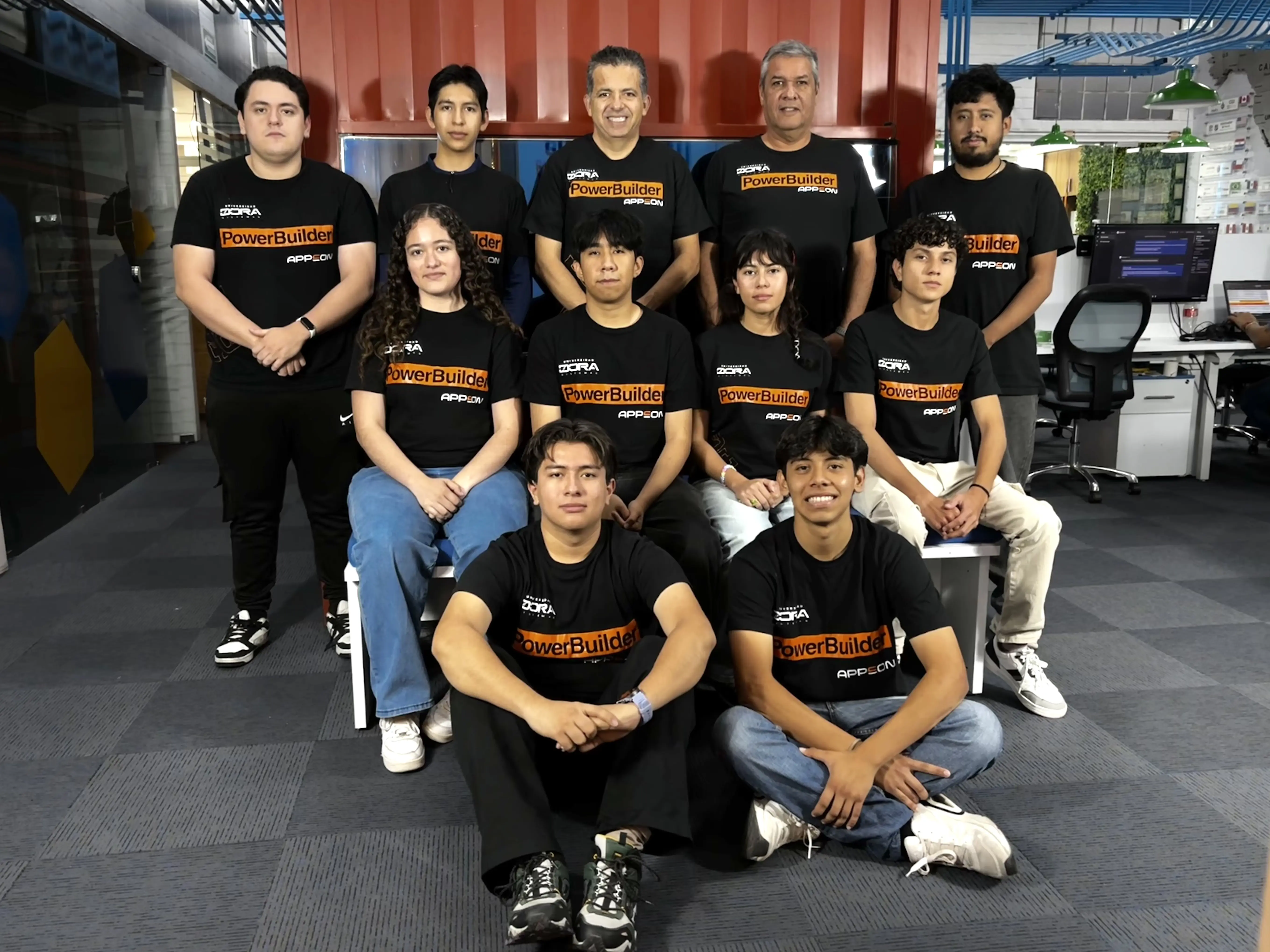 Un grupo de once personas, en su mayoría jóvenes adultos y dos hombres mayores, posan en el interior en tres filas con camisetas negras a juego de PowerBuilder; algunos están sentados y otros de pie, en un entorno de oficina moderno con escritorios, sillas y ordenadores al fondo.