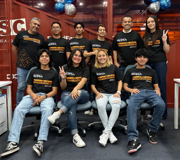 Un grupo de diez personas que pertenecen a la segunda generación dora, posando en el interior, ocho de pie y dos sentadas, todas con camisetas negras a juego en las que se lee PowerBuilder, en una habitación decorada con globos azules y plateados.