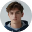 Portrait of a young man with short brown hair wearing a dark hoodie against a plain background.