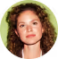 Portrait of a young woman with curly hair wearing a black top, smiling slightly against a green blurred leafy background.