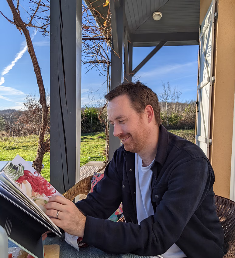 Man with short brown hair and beard sitting outdoors at a table, smiling while looking at a colorful book.