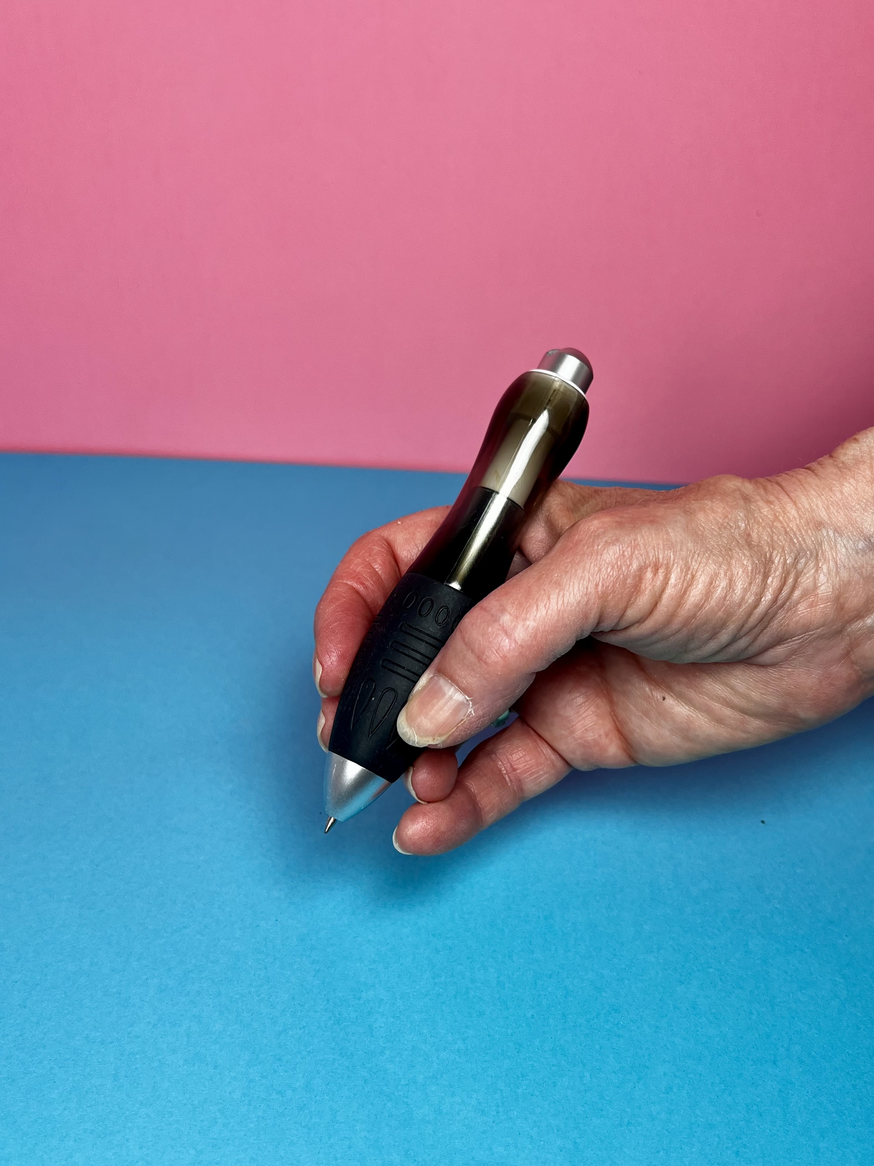 Hand holding a weighted pen with a black grip on a blue and pink background.