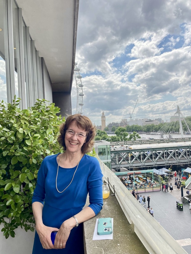 Smiling woman in blue dress standing on a balcony with London Eye and Big Ben in the background.