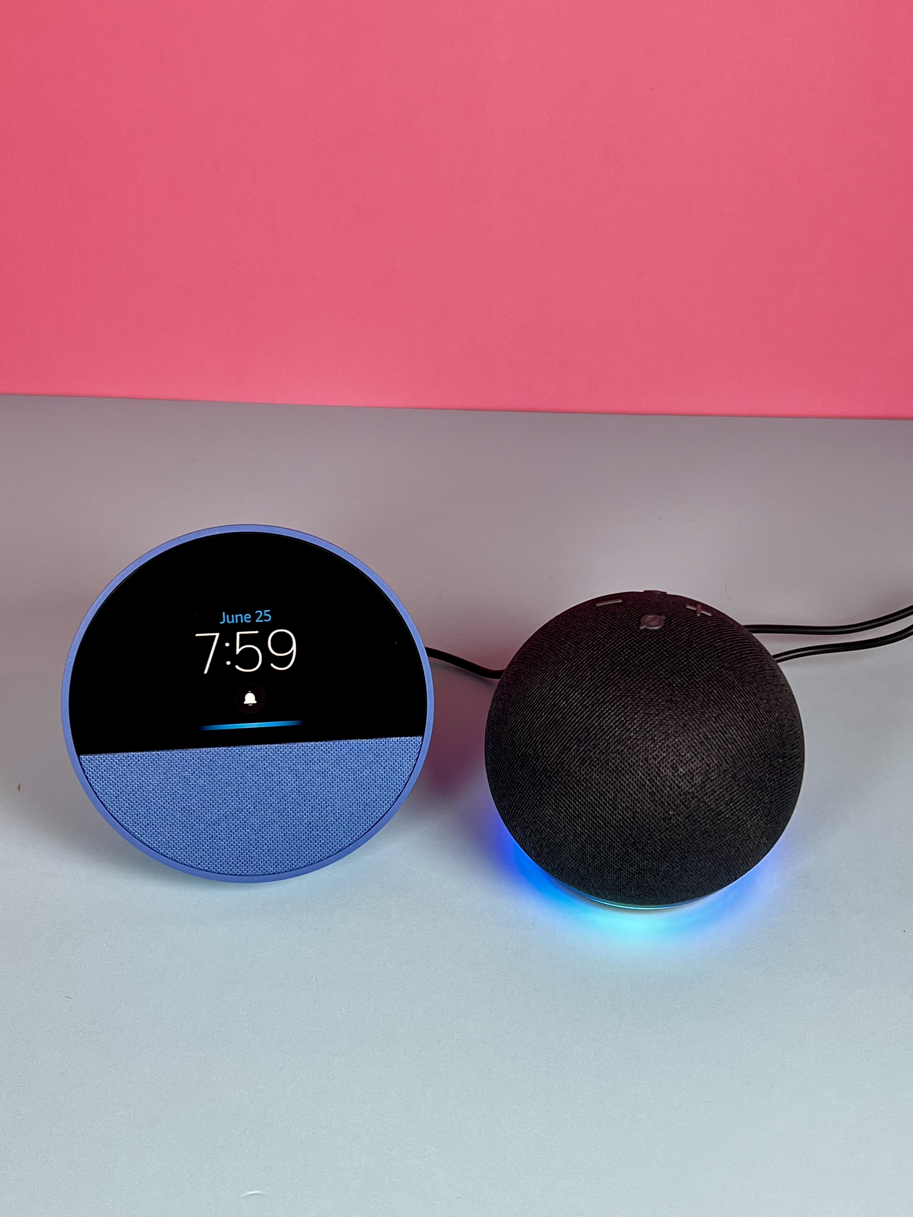 Two smart speakers on a surface: a blue Echo Spot showing 7:59 and a black Echo Dot with blue light.