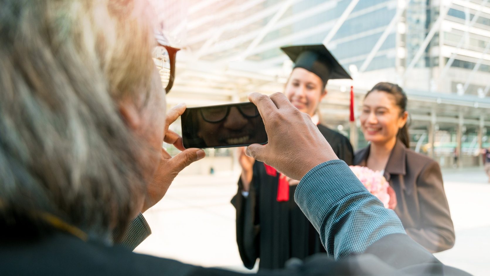 A visiting father takes a smartphone photo of a graduate in cap and gown and her mother on a campus plaza.