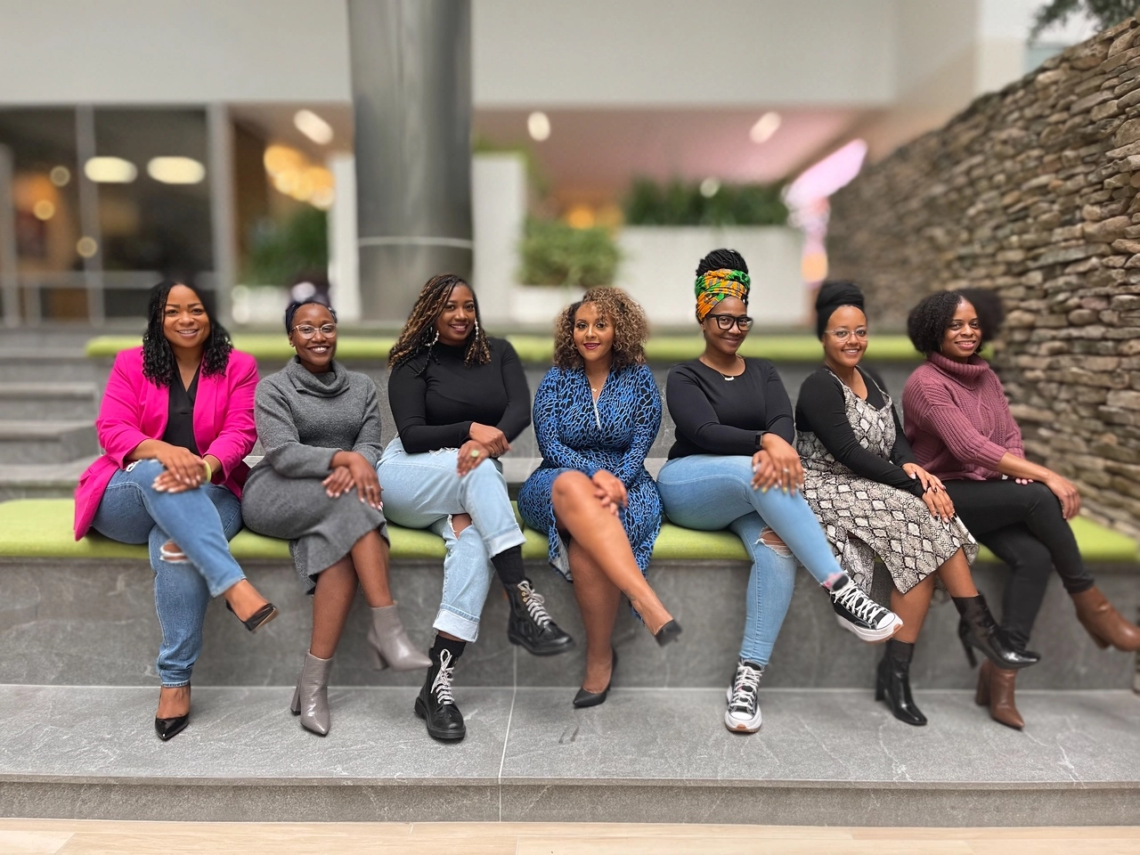 Seven women sitting on a green bench indoors, smiling and posing with crossed legs.