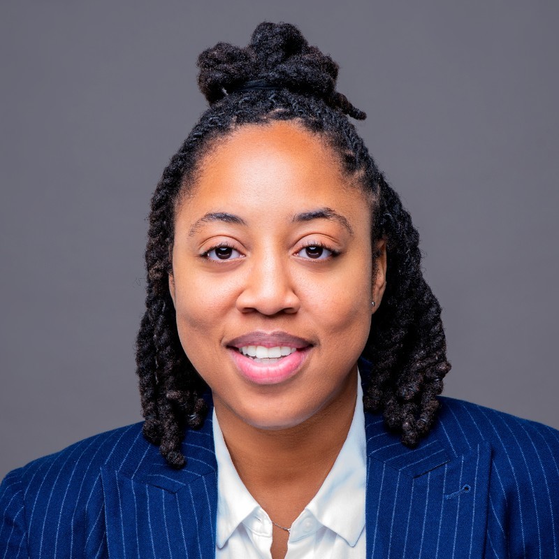 Smiling woman with braided hair wearing a pinstripe blue blazer and white shirt on gray background.