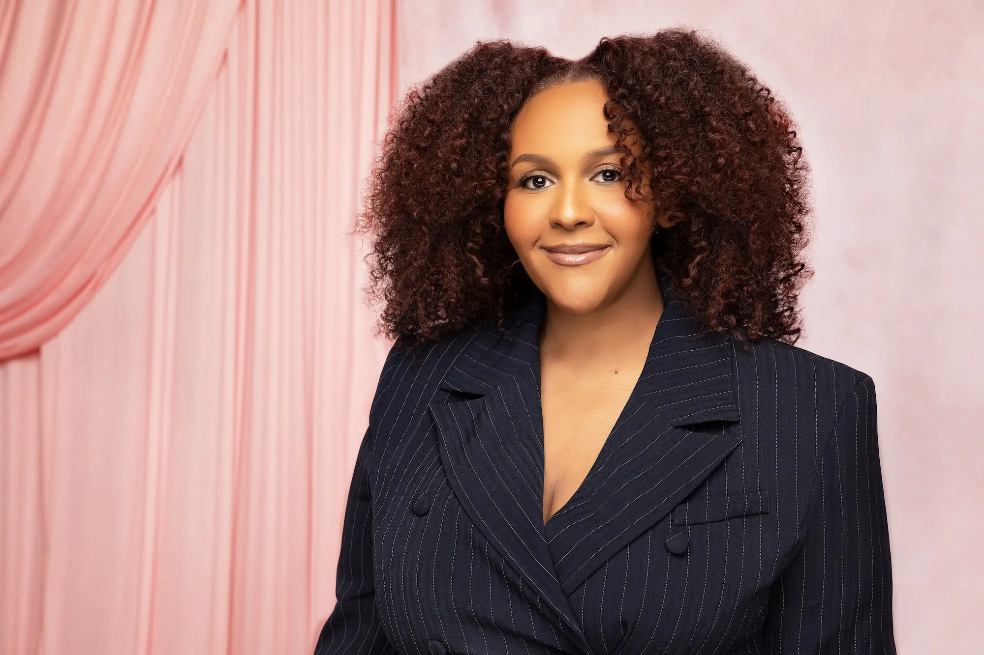 Smiling woman with voluminous curly hair wearing a dark pinstripe blazer against a pink backdrop.