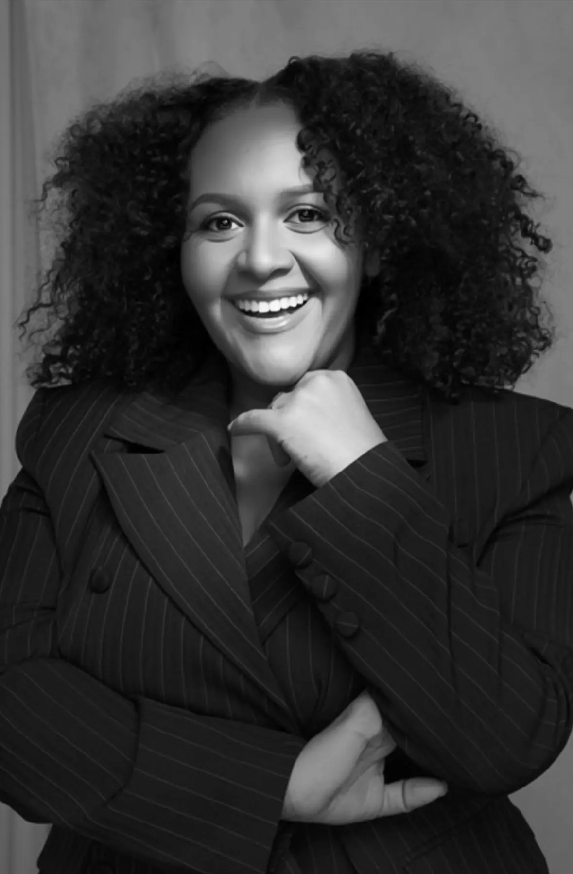 Smiling woman with curly hair wearing a pinstripe blazer, resting her chin on her hand.