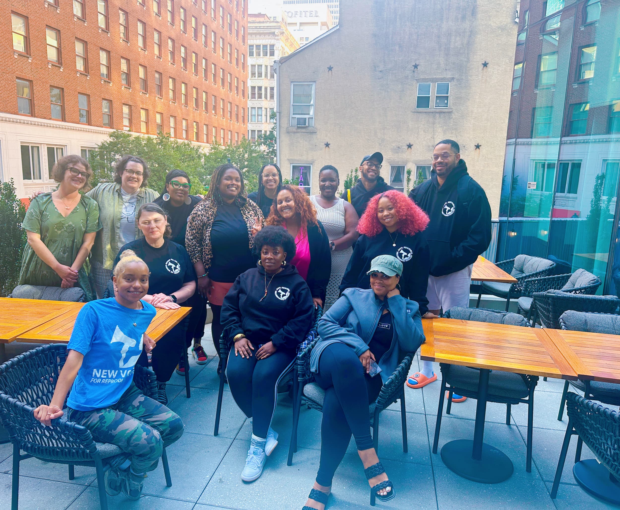 Group of smiling people posing outdoors on a patio with chairs and tables, city buildings in background.