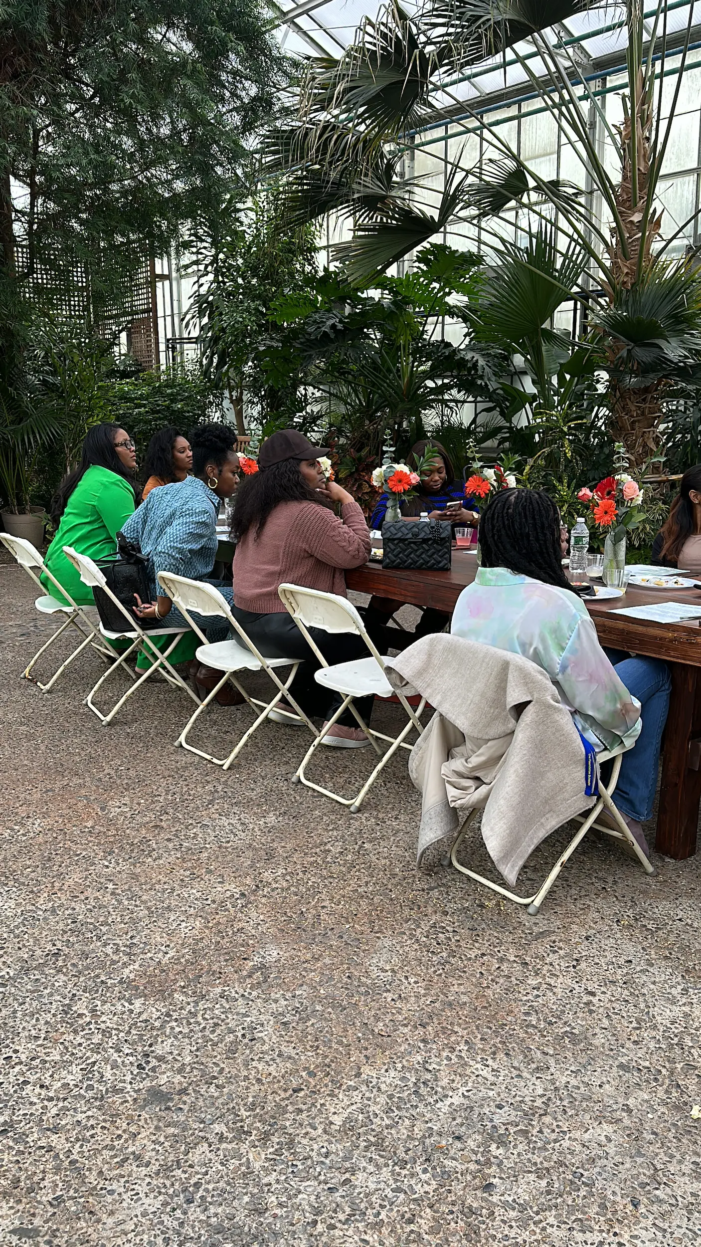 Group of women sitting on white folding chairs around a wooden table with flowers in a greenhouse.