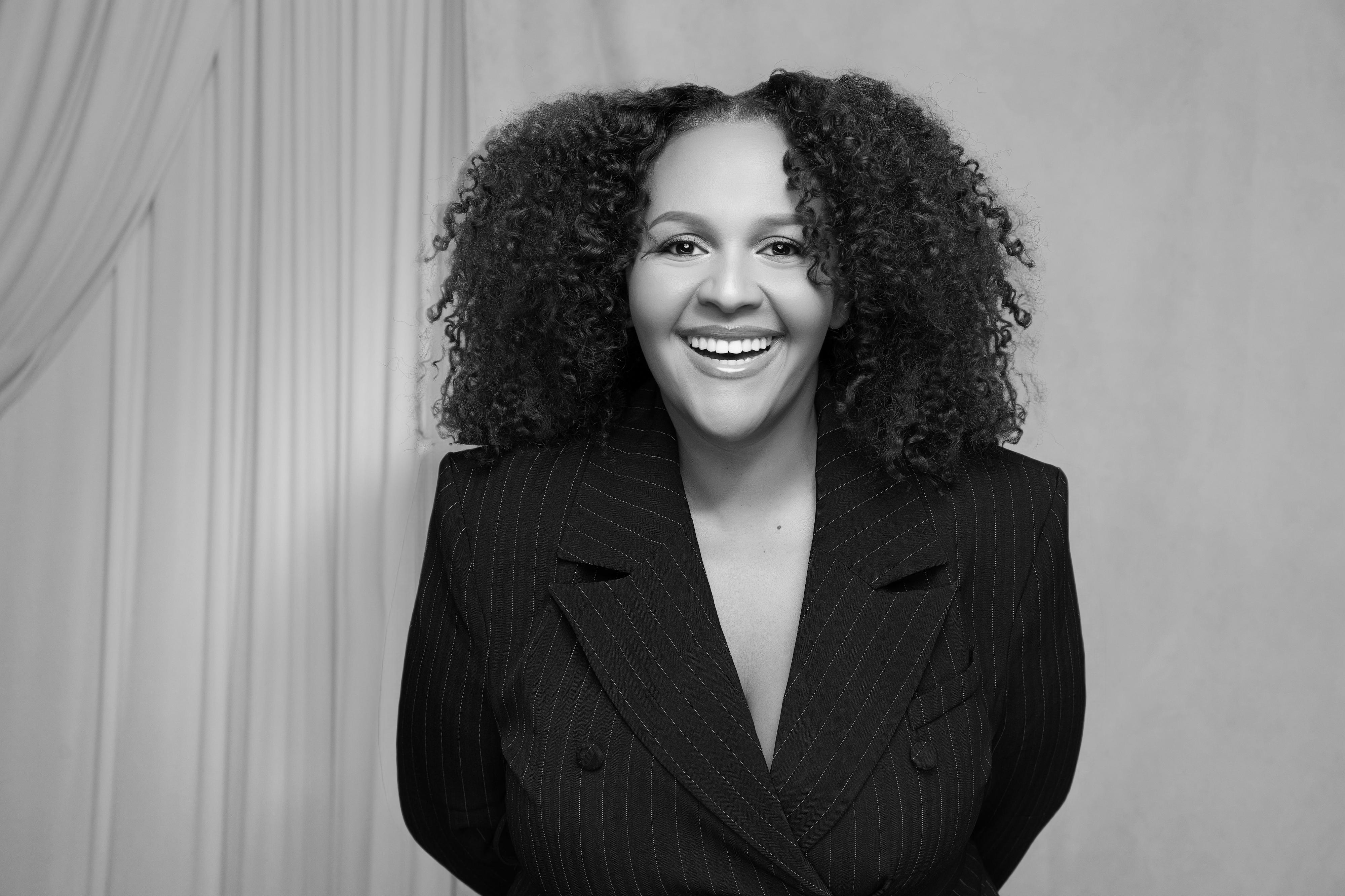 A woman with curly hair smiling widely in a pinstripe blazer against a light backdrop.