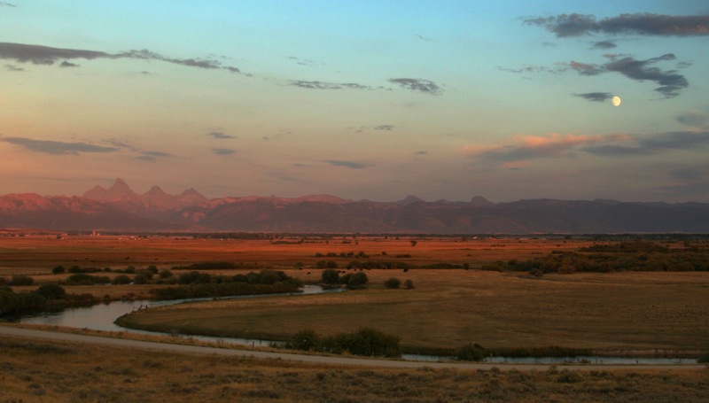 Wide valley with a small pond in the foreground, winding dirt road, scattered houses, and a forested mountain with ski slopes in the background.