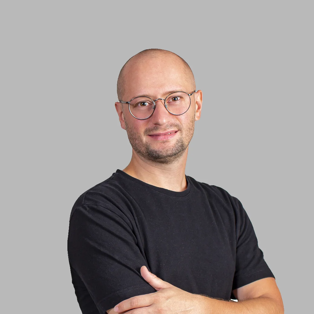 Bald man with round glasses wearing a black t-shirt, smiling with arms crossed on a gray background.