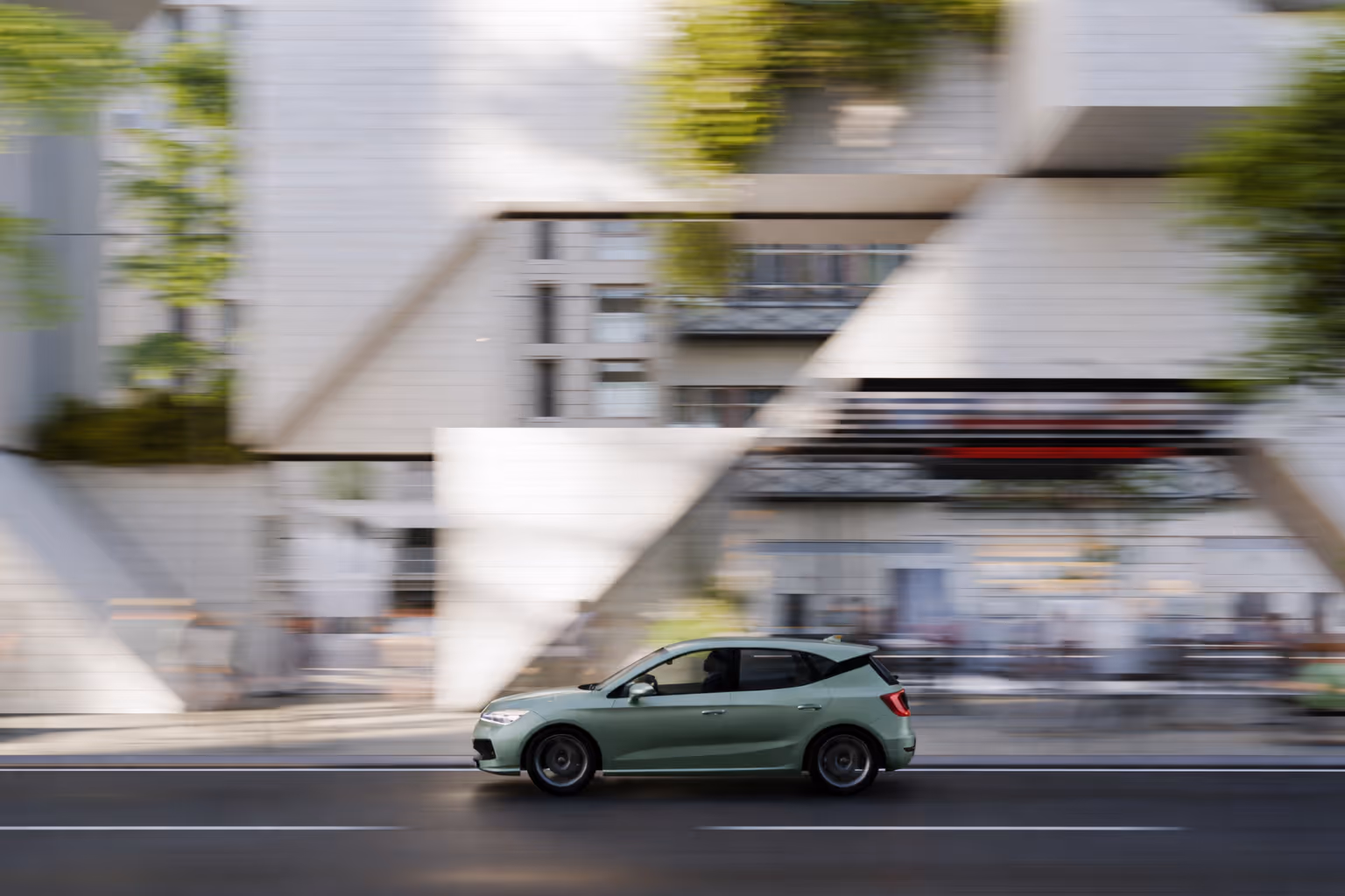 Green compact car driving on a city street with blurred buildings and trees in the background.