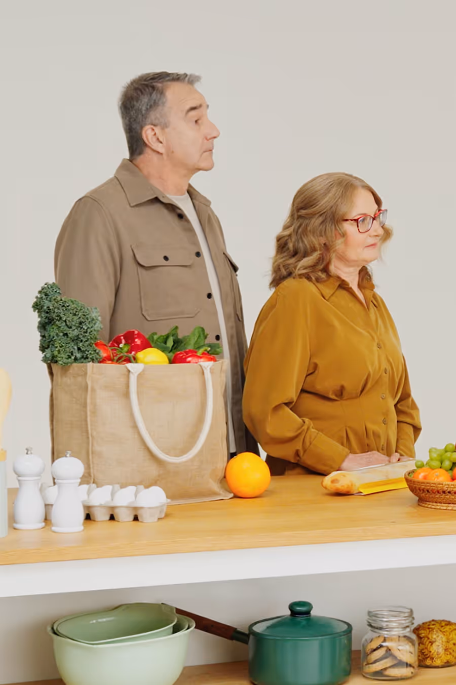 Mature couple standing at a kitchen counter with groceries including leafy greens, bell peppers, eggs, an orange, and a basket of fruit.