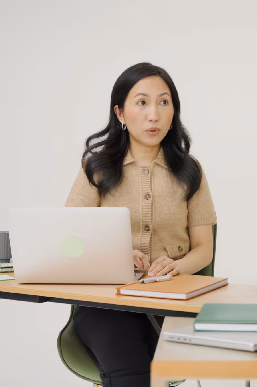 Woman with long dark hair wearing a tan buttoned sweater sitting at a desk with a laptop and notebooks.