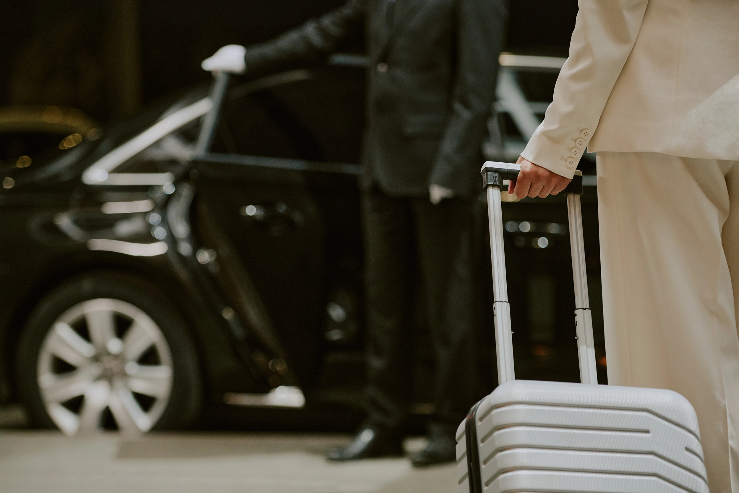 Person in a light-colored suit holding a suitcase handle in front of a black car with a chauffeur opening the door.