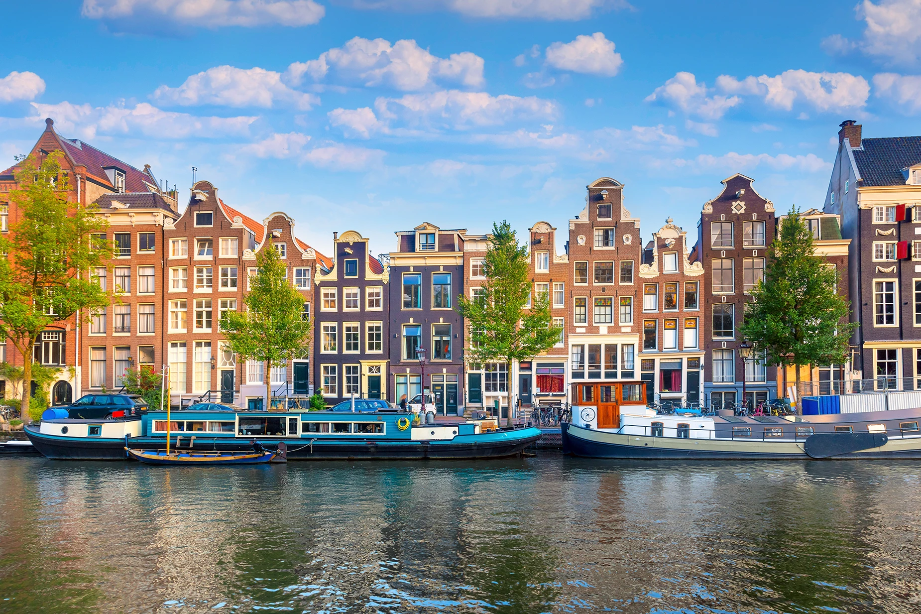 Colorful traditional Dutch canal houses along a waterway with boats moored in front under a partly cloudy sky.